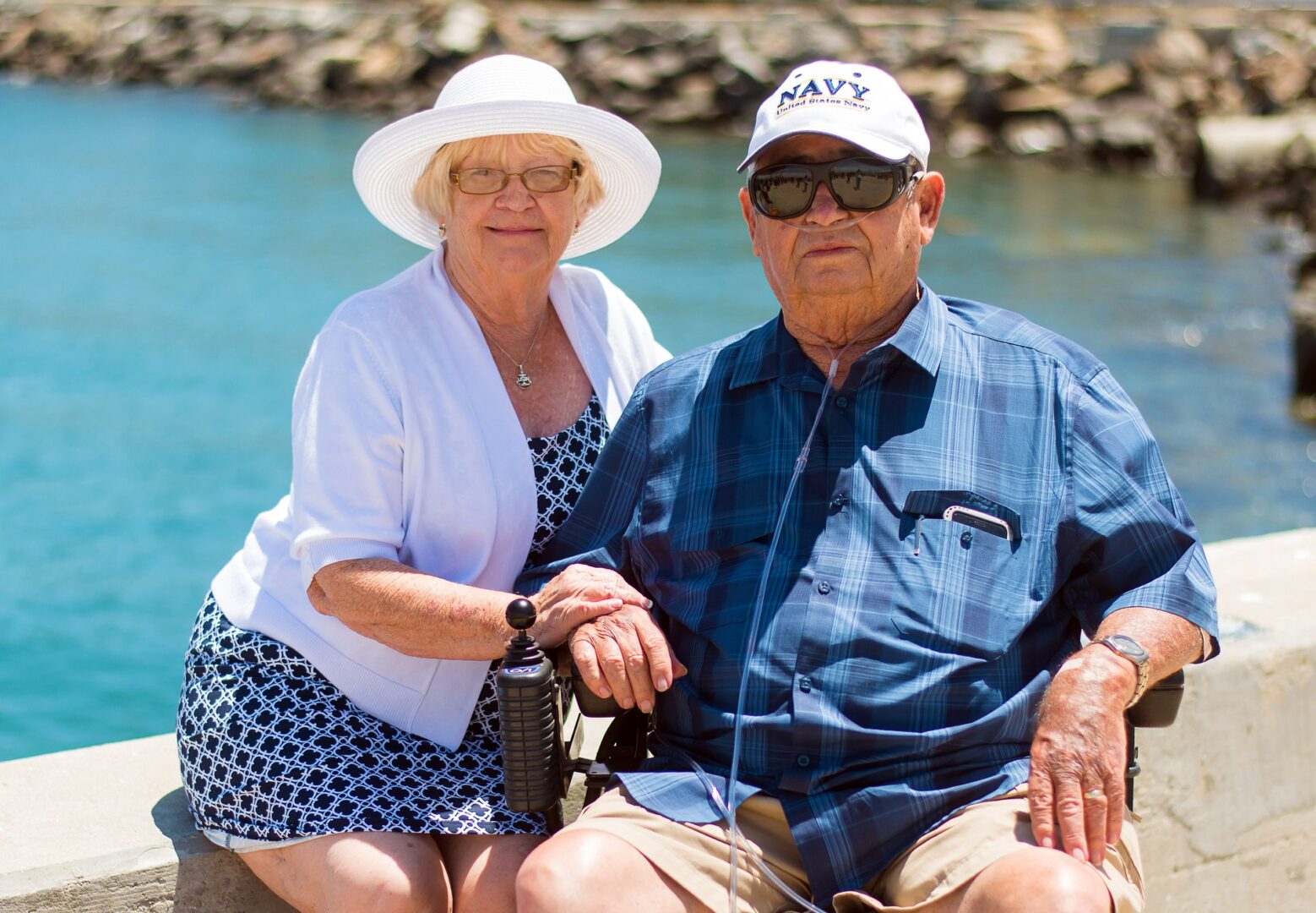 older couple sitting by the water.  Man has an oxygen cannula in his nose and a NAVY cap and is sitting in a wheel chair.