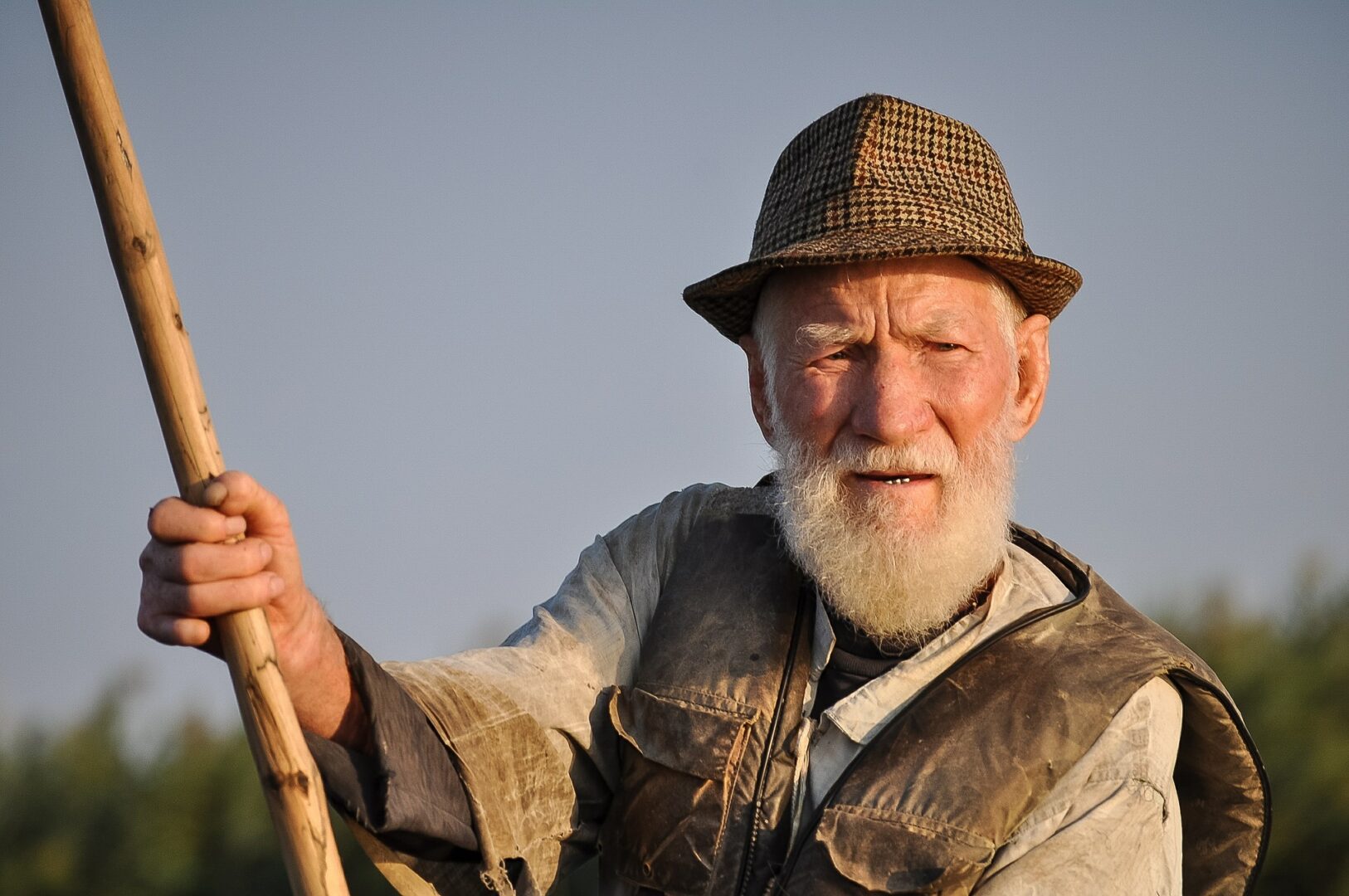 Man with white beard and hat rowing through what looks like a swamp