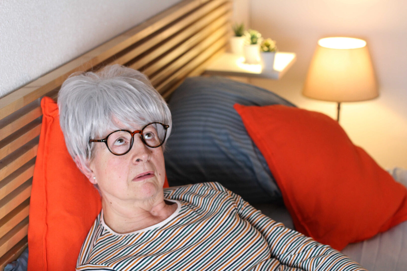 Woman with Gray hair and glasses lying in bed and looking up at the sky as if in despair.